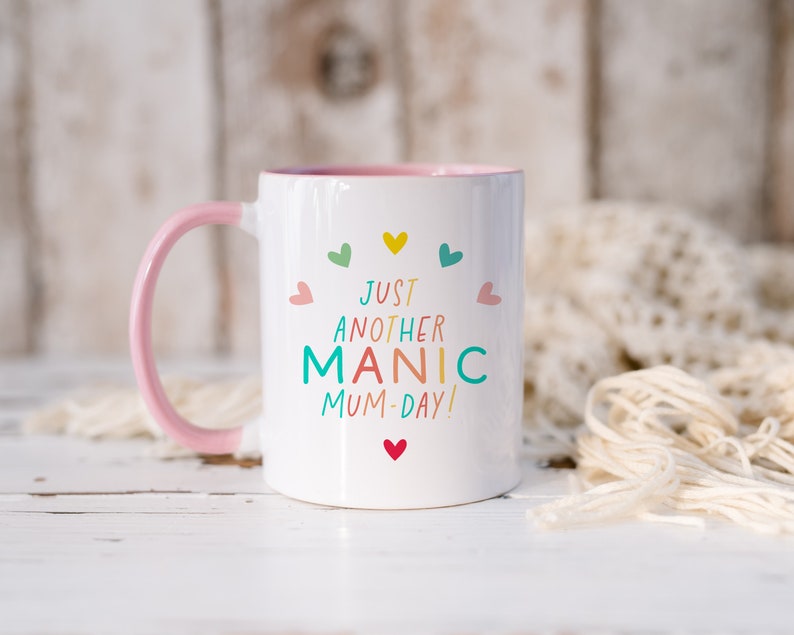 May include: A white ceramic mug with a light pink handle and interior rim. The front features the colorful text "JUST ANOTHER MANIC MUM-DAY!" surrounded by small, multi-colored hearts in shades of yellow, teal, green, and pink.