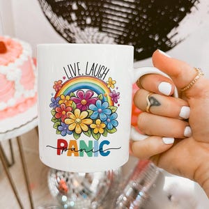 Può includere: Tazza in ceramica bianca con un design a arcobaleno e fiori. Il testo "Live, Laugh, Panic" è stampato sulla tazza.