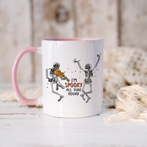 May include: White ceramic mug with a pink rim and handle. The mug features a black and white illustration of two skeletons, one playing a violin. The text "I'm Spooky All Year Round" is printed on the mug.