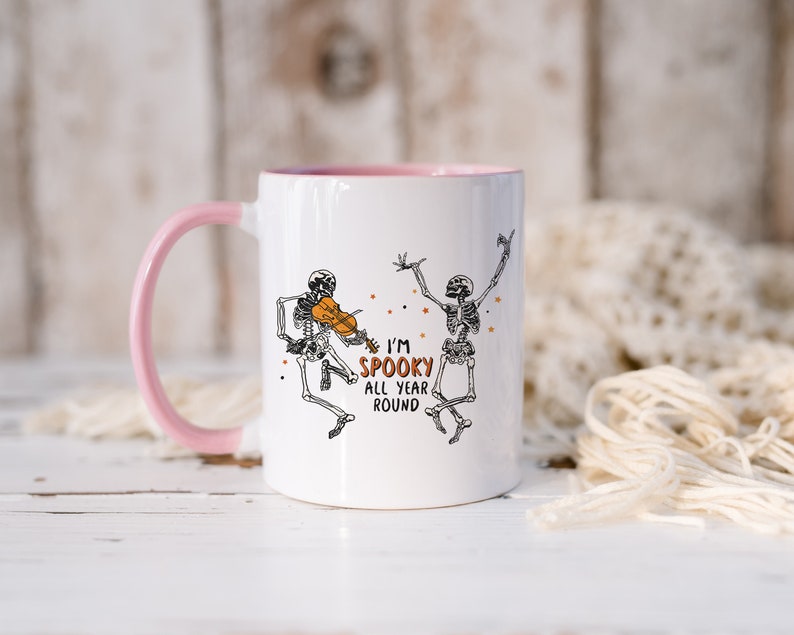 May include: White ceramic mug with a pink rim and handle. The mug features a black and white illustration of two skeletons, one playing a violin. The text "I'm Spooky All Year Round" is printed on the mug.