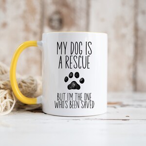 May include: White ceramic mug with a yellow handle. The mug has black text that reads "My dog is a rescue but I'm the one who's been saved". A black paw print is in the middle of the text.