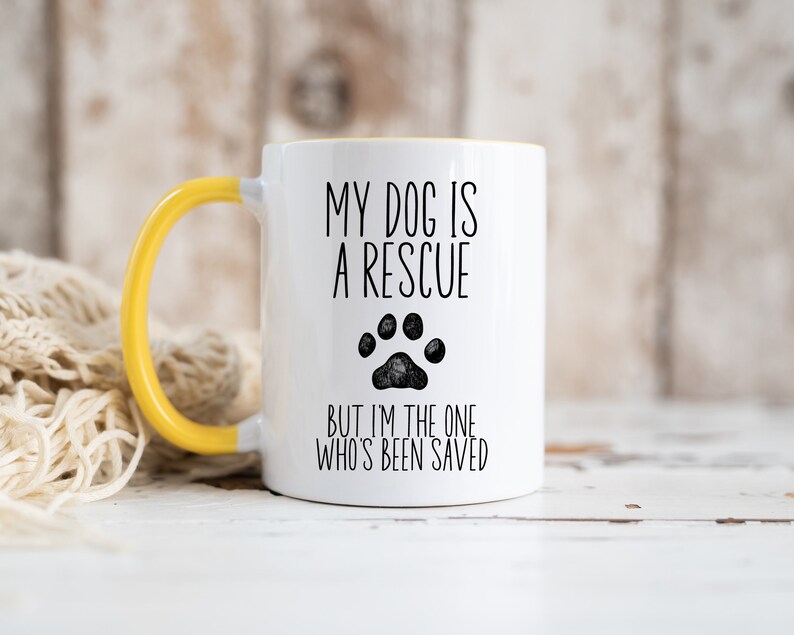May include: White ceramic mug with a yellow handle. The mug has black text that reads "My dog is a rescue but I'm the one who's been saved". A black paw print is in the middle of the text.