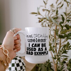 May include: White ceramic mug with the text "I'm not totally useless I can be used as a bad example" in black. The mug has a large handle and is held by a person. The background includes greenery and a black and white checkered pattern.