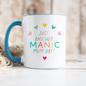 May include: A white ceramic mug with a blue handle and interior rim. The front features the colorful text "JUST ANOTHER MANIC MUM-DAY!" surrounded by small, multi-colored heart icons in shades of pink, yellow, teal, and red, arranged in a circular pattern.