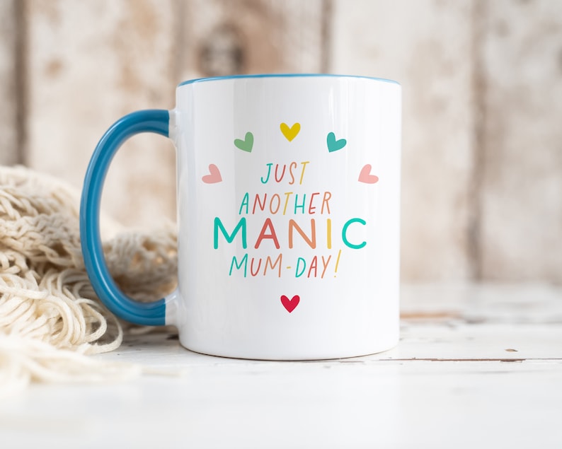May include: A white ceramic mug with a blue handle and interior rim. The front features the colorful text "JUST ANOTHER MANIC MUM-DAY!" surrounded by small, multi-colored heart icons in shades of pink, yellow, teal, and red, arranged in a circular pattern.