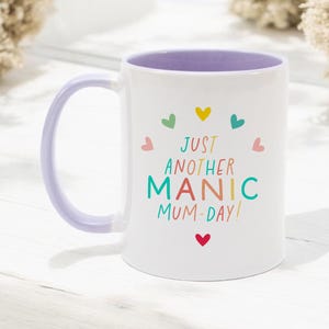 May include: A white ceramic mug with a light purple handle and interior. The front features the text "JUST ANOTHER MANIC MUM-DAY!" in colorful letters, surrounded by a circular arrangement of small, multi-colored hearts in red, yellow, teal, and pink.