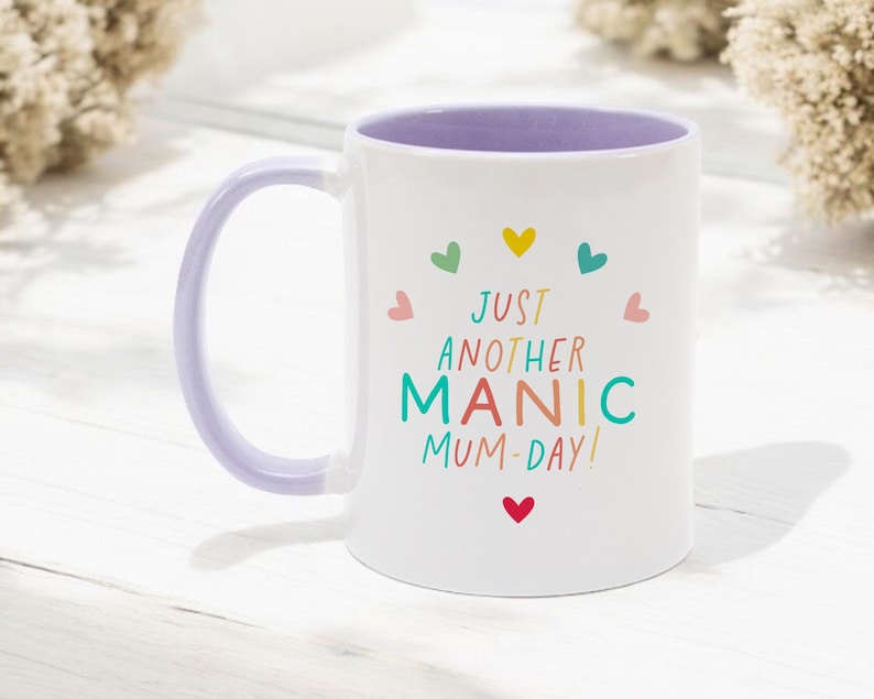May include: A white ceramic mug with a light purple handle and interior. The front features the text "JUST ANOTHER MANIC MUM-DAY!" in colorful letters, surrounded by a circular arrangement of small, multi-colored hearts in red, yellow, teal, and pink.