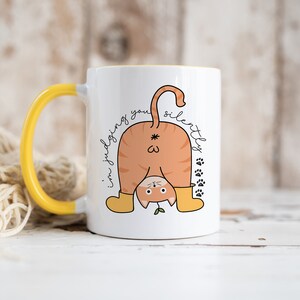 May include: White ceramic mug with a yellow handle. The mug features a cartoon illustration of a cat upside down with the text "I'm judging you silently".