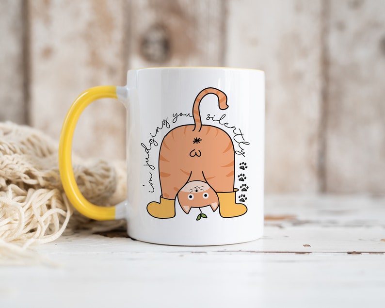May include: White ceramic mug with a yellow handle. The mug features a cartoon illustration of a cat upside down with the text "I'm judging you silently".