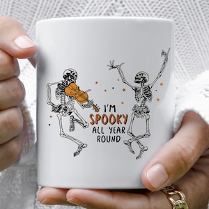 May include: White ceramic coffee mug with a black and white illustration of two skeletons dancing. One skeleton is playing a violin. The text "I'm Spooky All Year Round" is printed on the mug.