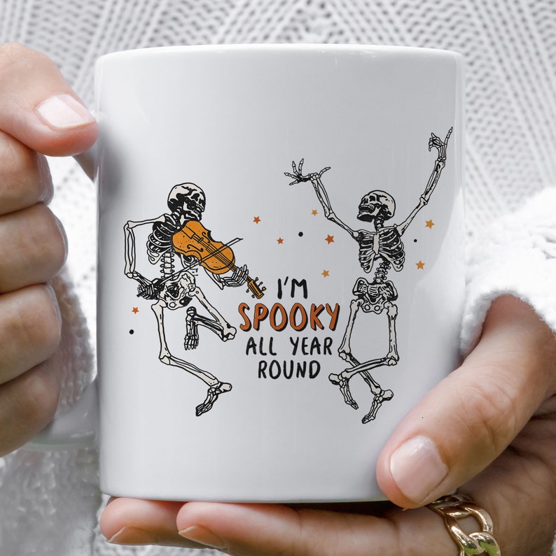 May include: White ceramic coffee mug with a black and white illustration of two skeletons dancing. One skeleton is playing a violin. The text "I'm Spooky All Year Round" is printed on the mug.