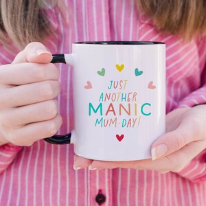 May include: A white ceramic mug with a black handle and interior rim. The front features the text "JUST ANOTHER MANIC MUM-DAY!" in multi-colored letters, surrounded by small, colorful heart icons in shades of red, yellow, teal, and pink.