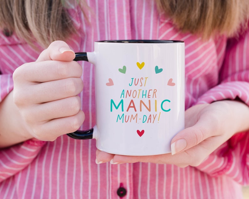 May include: A white ceramic mug with a black handle and interior rim. The front features the text "JUST ANOTHER MANIC MUM-DAY!" in multi-colored letters, surrounded by small, colorful heart icons in shades of red, yellow, teal, and pink.