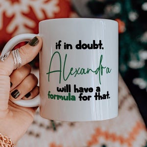 Puede incluir: Taza de cerámica blanca con el texto "if in doubt, Alexandra will have a formula for that" en letras negras y verdes. La taza tiene un asa curva y la sostiene una persona.