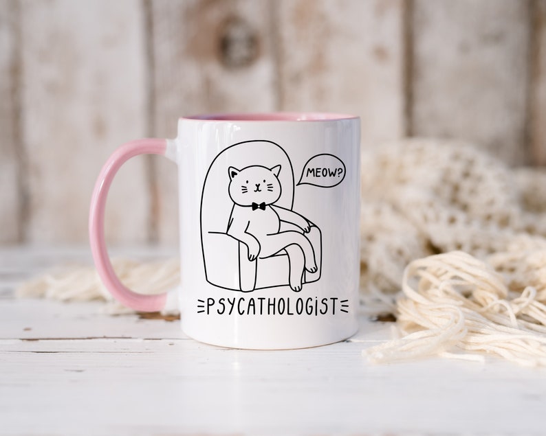 a white ceramic mug, with a pink handle, with an illustration of a cat, wearing a bowtie, sitting on an armchair. Beside the cat is a speech bubble with the text 'Meow?' - with text below the graphic stating 'Psycathologist'.