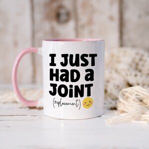 May include: A white ceramic mug with a light pink handle and interior rim. The front features bold black text that reads I JUST HAD A JOINT (replacement) followed by a small yellow winking smiley face icon with pink cheeks.