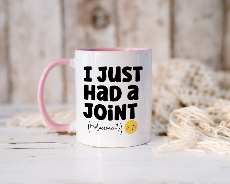 May include: A white ceramic mug with a light pink handle and interior rim. The front features bold black text that reads I JUST HAD A JOINT (replacement) followed by a small yellow winking smiley face icon with pink cheeks.