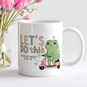 Può includere: Una tazza in ceramica bianca con una rana verde che guida uno scooter rosa. La tazza ha la scritta "Let's do this before my anxiety kicks in." stampata su di essa.