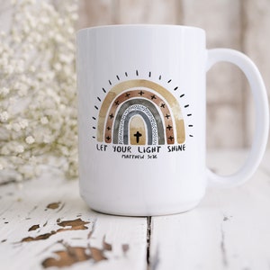 May include: White ceramic coffee mug featuring a rainbow design in shades of brown, grey, and gold. The rainbow has a cross in the centre and the text "LET YOUR LIGHT SHINE" with "MATTHEW 5:16" below.
