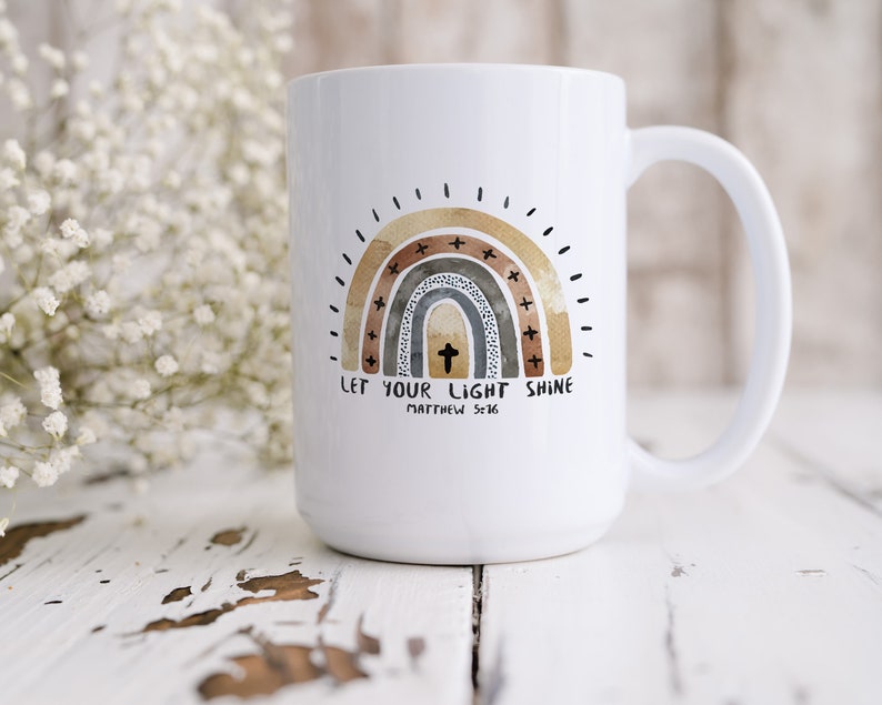 May include: White ceramic coffee mug featuring a rainbow design in shades of brown, grey, and gold. The rainbow has a cross in the centre and the text "LET YOUR LIGHT SHINE" with "MATTHEW 5:16" below.