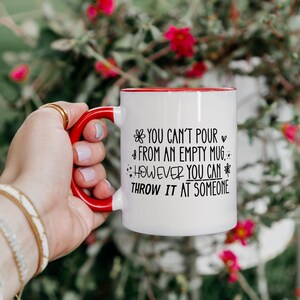 May include: White ceramic mug with a red handle and rim. The mug features black text that reads "YOU CAN'T POUR FROM AN EMPTY MUG. HOWEVER YOU CAN THROW IT AT SOMEONE."