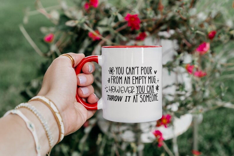 May include: White ceramic mug with a red handle and rim. The mug features black text that reads "YOU CAN'T POUR FROM AN EMPTY MUG. HOWEVER YOU CAN THROW IT AT SOMEONE."