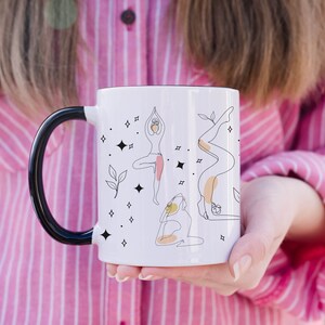 May include: White ceramic mug with a black handle and rim, decorated with line art illustrations of yoga poses, stars, and leaves. The mug is held by a person wearing a pink and white striped shirt.