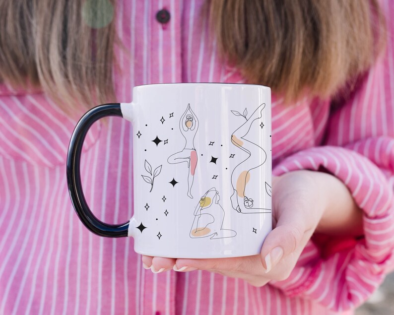 May include: White ceramic mug with a black handle and rim, decorated with line art illustrations of yoga poses, stars, and leaves. The mug is held by a person wearing a pink and white striped shirt.