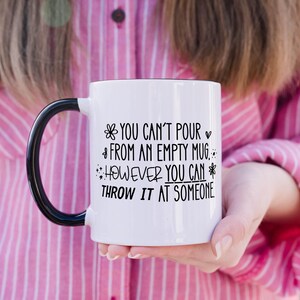 May include: White ceramic mug with a black handle and rim. The mug features the text "YOU CAN'T POUR FROM AN EMPTY MUG, HOWEVER YOU CAN THROW IT AT SOMEONE" in black lettering. The mug is held by a person wearing a pink shirt.