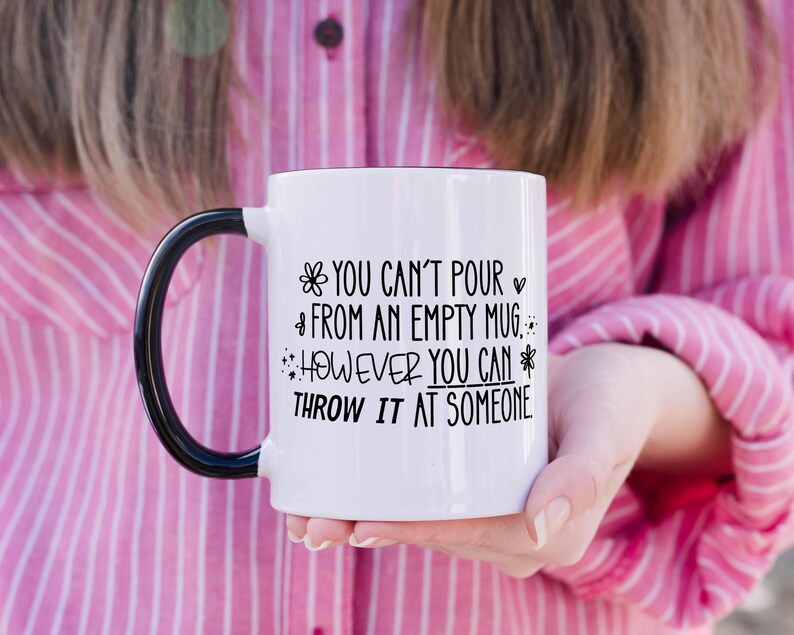 May include: White ceramic mug with a black handle and rim. The mug features the text "YOU CAN'T POUR FROM AN EMPTY MUG, HOWEVER YOU CAN THROW IT AT SOMEONE" in black lettering. The mug is held by a person wearing a pink shirt.