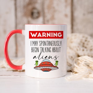 Op de afbeelding: Een witte koffiemok met een rode rand en handvat. De mok heeft een zwart-witte afbeelding met de tekst "WARNING I MAY SPONTANEOUSLY BEGIN TALKING ABOUT ALIENS" en twee groene aliens die naast een rode vliegende schotel staan.