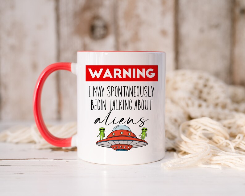 Op de afbeelding: Een witte koffiemok met een rode rand en handvat. De mok heeft een zwart-witte afbeelding met de tekst "WARNING I MAY SPONTANEOUSLY BEGIN TALKING ABOUT ALIENS" en twee groene aliens die naast een rode vliegende schotel staan.