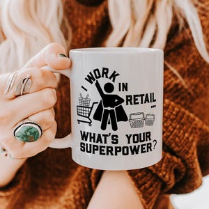 Könnte beinhalten: Weiße Keramik-Tasse mit dem schwarzen Text "I WORK IN RETAIL WHAT'S YOUR SUPERPOWER?". Das Design zeigt eine Strichmännchen-Superheldenfigur mit Einkaufswagen und Körben.