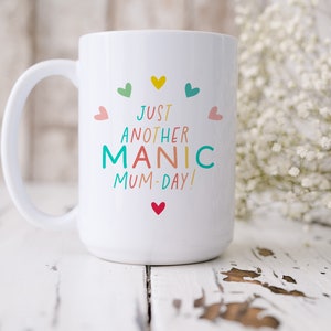 May include: A white ceramic mug featuring the colorful text JUST ANOTHER MANIC MUM-DAY! printed in the center. The design is surrounded by small, multi-colored hearts in shades of yellow, teal, pink, and red, arranged in a circular pattern.