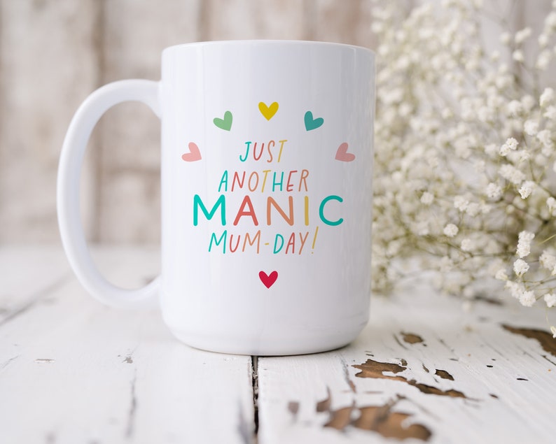 May include: A white ceramic mug featuring the colorful text JUST ANOTHER MANIC MUM-DAY! printed in the center. The design is surrounded by small, multi-colored hearts in shades of yellow, teal, pink, and red, arranged in a circular pattern.