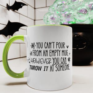 May include: White ceramic mug with a green handle and rim. The mug features the text "YOU CAN'T POUR FROM AN EMPTY MUG, HOWEVER YOU CAN THROW IT AT SOMEONE" in black lettering. Decorative bats are in the background.