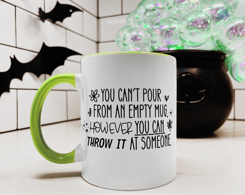 May include: White ceramic mug with a green handle and rim. The mug features the text "YOU CAN'T POUR FROM AN EMPTY MUG, HOWEVER YOU CAN THROW IT AT SOMEONE" in black lettering. Decorative bats are in the background.