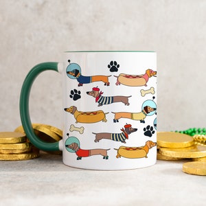 May include: White ceramic mug with a green handle featuring a repeating pattern of dachshunds in various costumes, including a space suit, a beret, and a hot dog. The dachshunds are all facing right and have black noses and brown eyes.