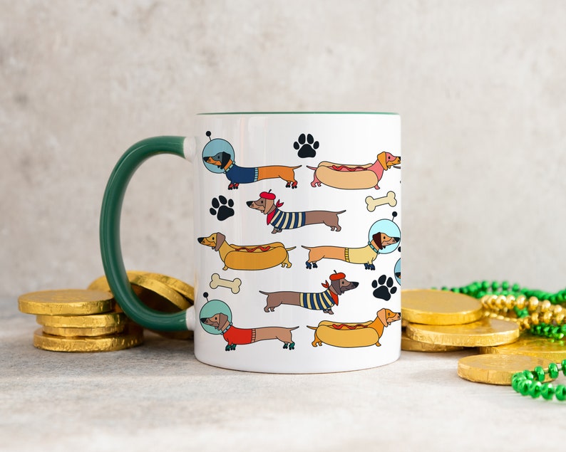 May include: White ceramic mug with a green handle featuring a repeating pattern of dachshunds in various costumes, including a space suit, a beret, and a hot dog. The dachshunds are all facing right and have black noses and brown eyes.