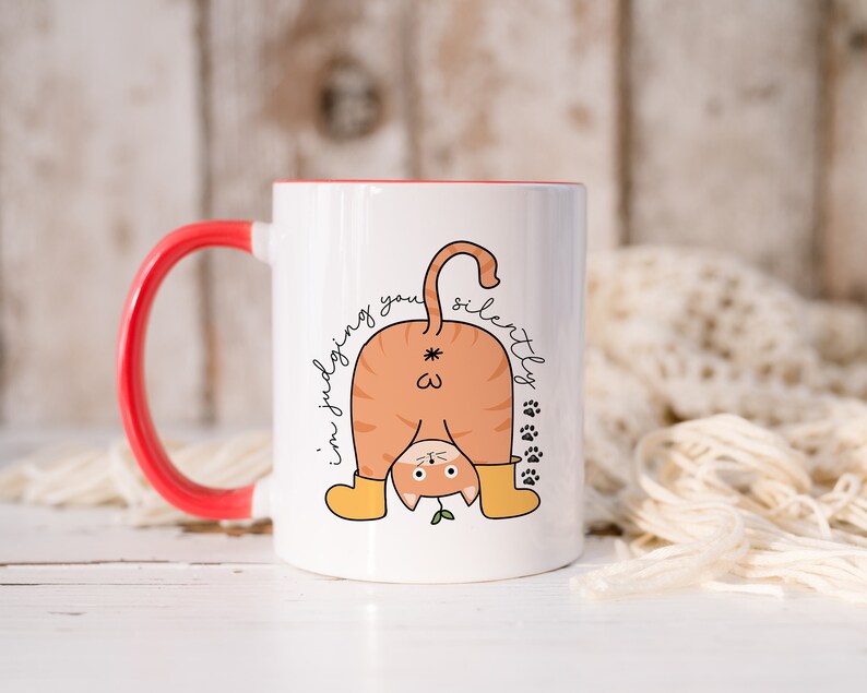 May include: White ceramic mug with a red handle. The mug features a cartoon illustration of a cat with its back turned, looking over its shoulder with the text "I'm judging you silently".