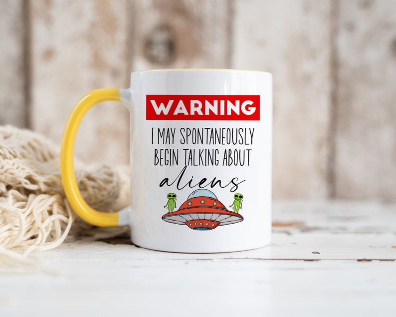 Op de afbeelding: Witte keramische mok met een gele handgreep. De mok heeft een rood waarschuwingsbord met de tekst "WAARSCHUWING IK KAN SPONTAN BEGINNEN OVER BUITENAARDSE WEZENS TE PRATEN". Aan weerszijden van de tekst staan twee groene buitenaardse wezens in vliegende schotels.