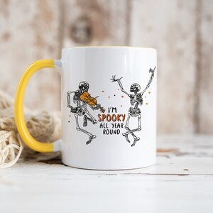 May include: White ceramic mug with a yellow handle. The mug features a black and white illustration of two skeletons, one playing a violin and the other dancing. The text "I'm Spooky All Year Round" is printed in orange on the mug.
