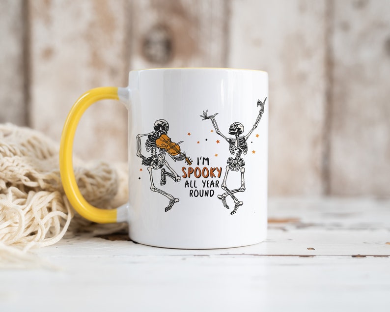 May include: White ceramic mug with a yellow handle. The mug features a black and white illustration of two skeletons, one playing a violin and the other dancing. The text "I'm Spooky All Year Round" is printed in orange on the mug.
