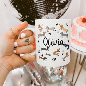 Pode incluir: Uma caneca de cer&acirc;mica branca com um padr&atilde;o repetitivo de c&atilde;es dachshund de v&aacute;rias cores, pequenos cora&ccedil;&otilde;es pretos e pegadas. O nome Olivia est&aacute; impresso numa fonte cursiva preta no centro da caneca, rodeado pelos c&atilde;es ilustrados.