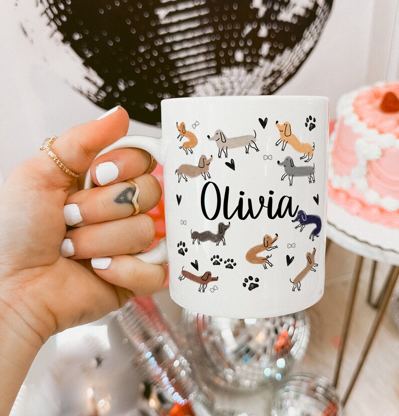 Pode incluir: Uma caneca de cer&acirc;mica branca com um padr&atilde;o repetitivo de c&atilde;es dachshund de v&aacute;rias cores, pequenos cora&ccedil;&otilde;es pretos e pegadas. O nome Olivia est&aacute; impresso numa fonte cursiva preta no centro da caneca, rodeado pelos c&atilde;es ilustrados.