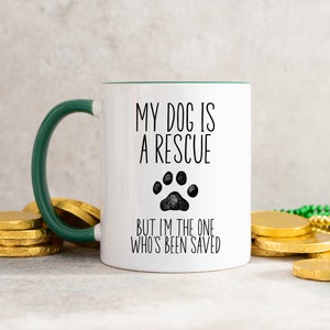 May include: White ceramic mug with a green rim and handle. The mug has black text that reads "My dog is a rescue but I'm the one who's been saved" with a black paw print graphic.