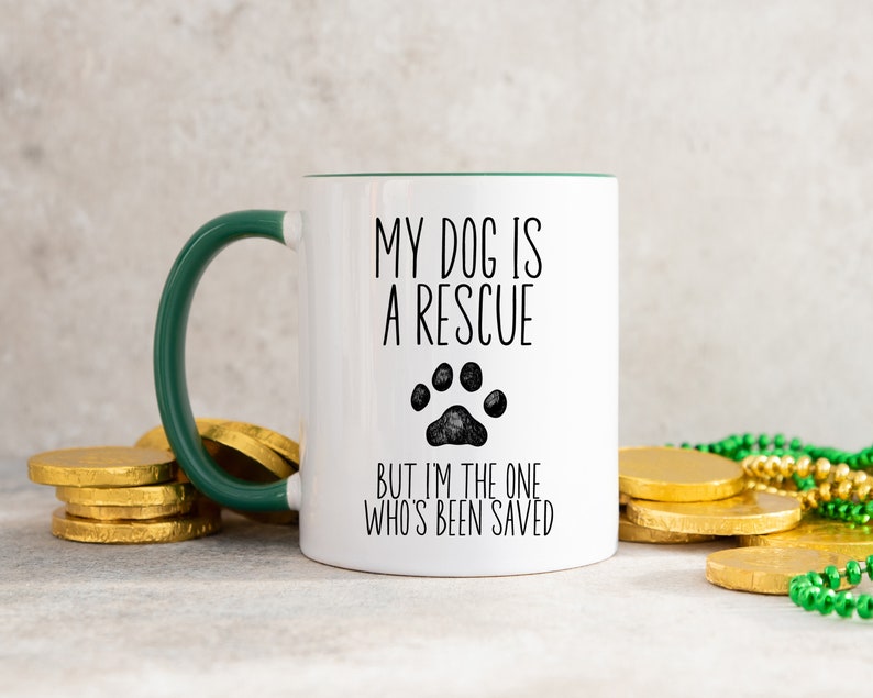 May include: White ceramic mug with a green rim and handle. The mug has black text that reads "My dog is a rescue but I'm the one who's been saved" with a black paw print graphic.