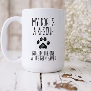 May include: White ceramic coffee mug with black text that reads "My dog is a rescue but I'm the one who's been saved". A black paw print is between the two lines of text.