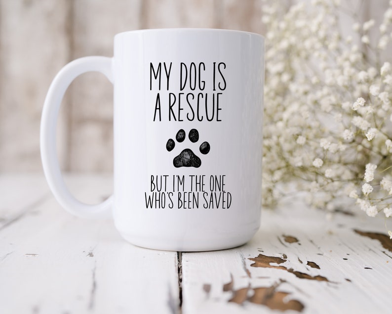 May include: White ceramic coffee mug with black text that reads "My dog is a rescue but I'm the one who's been saved". A black paw print is between the two lines of text.