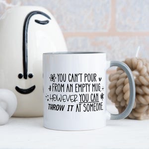 May include: White ceramic mug with a gray handle and rim, featuring the text "YOU CAN'T POUR FROM AN EMPTY MUG... HOWEVER YOU CAN THROW IT AT SOMEONE" in black. The mug is surrounded by decorative candles and a ceramic face.
